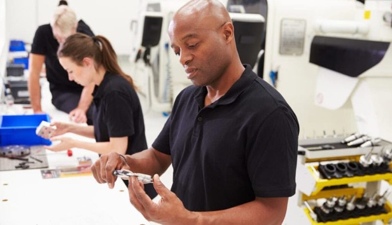 Man inspecting a metal part in a manufacturing workshop with workers in background.