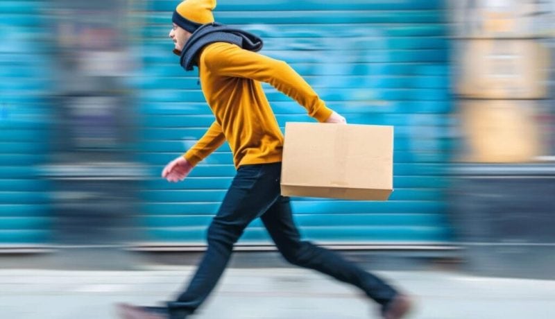 Fast-moving courier in yellow jacket carrying a box on city street.