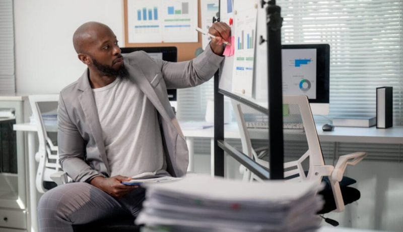 Businessman analyzing charts on digital screens in office with stacks of papers.