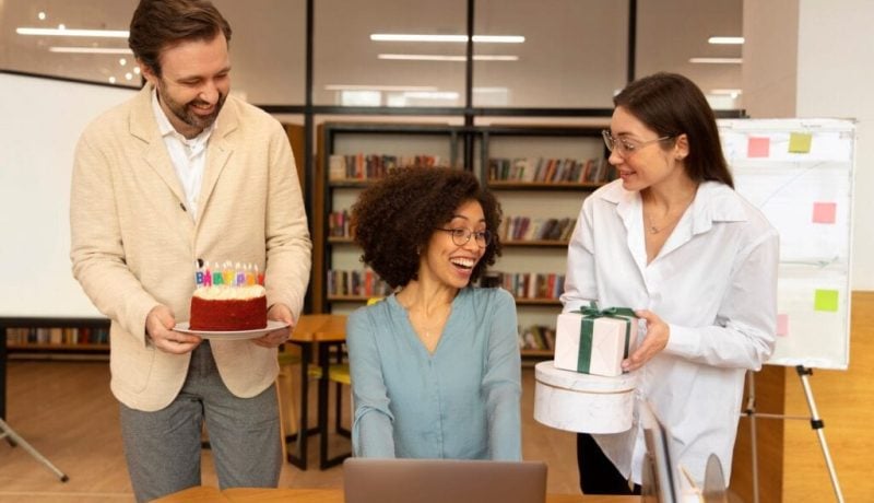 Happy colleagues celebrating with cake and gifts in an office setting.