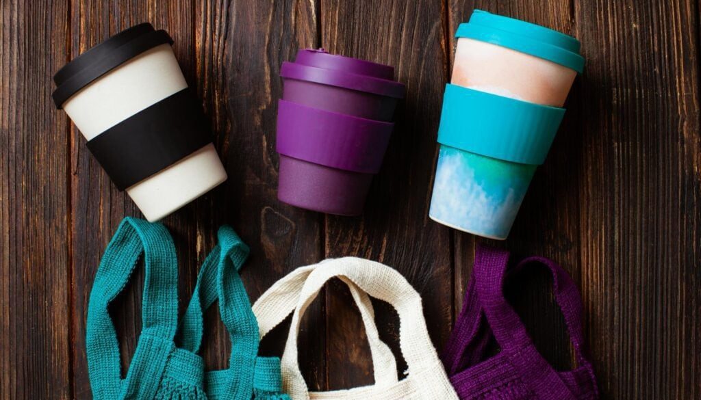 Colorful reusable coffee cups and fabric tote bags on wooden surface.