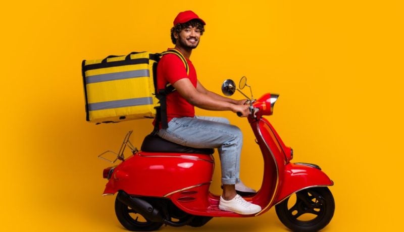 Courier wearing a red outfit delivering packages on a red scooter against a vibrant yellow background