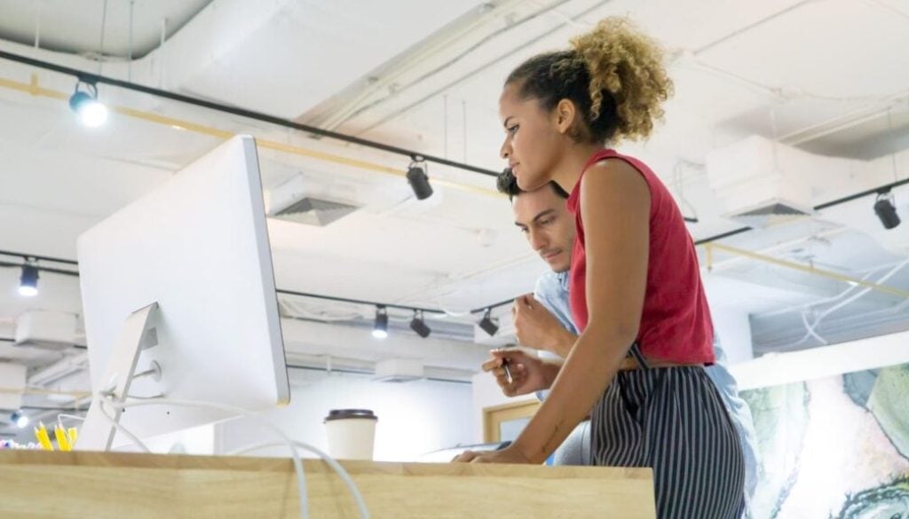 Two young professionals collaborating at a desk with a computer in a bright office.