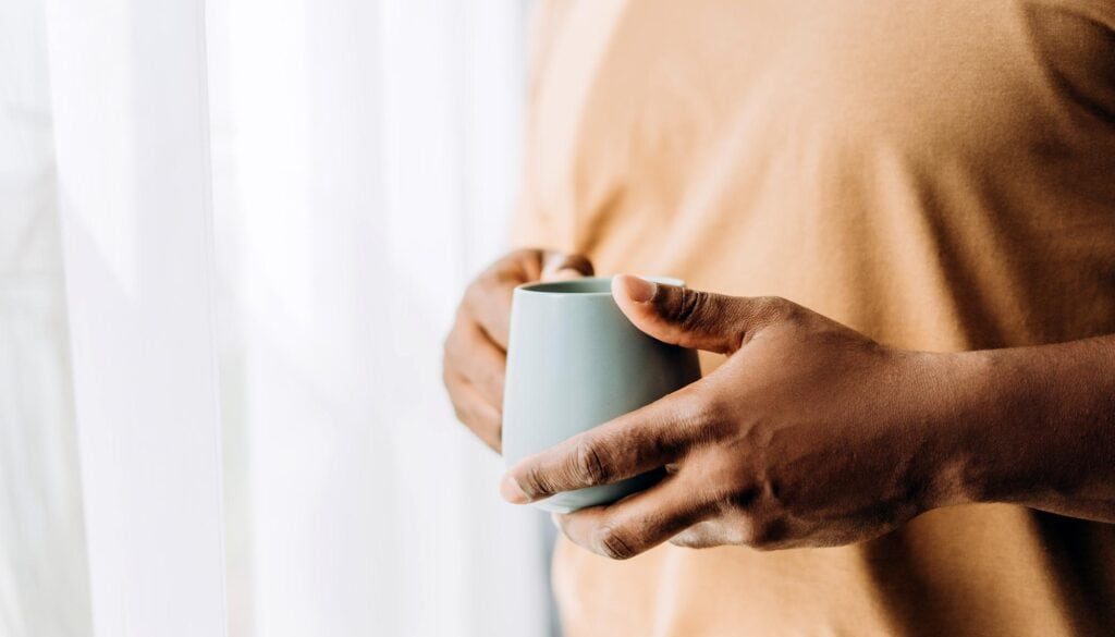 Person holding a coffee mug near a bright window, casual indoor setting.