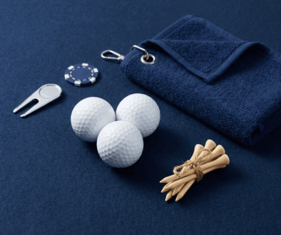 Three white golf balls with a navy towel, divot tool, ball markers, and tied tees on a dark blue surface -- a golf kit laid out neatly on the table.