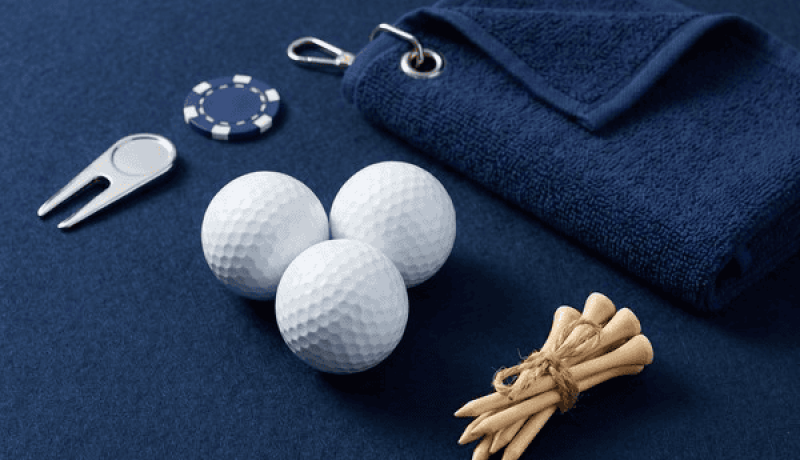 Three white golf balls with a navy towel, divot tool, ball markers, and tied tees on a dark blue surface -- a golf kit laid out neatly on the table.