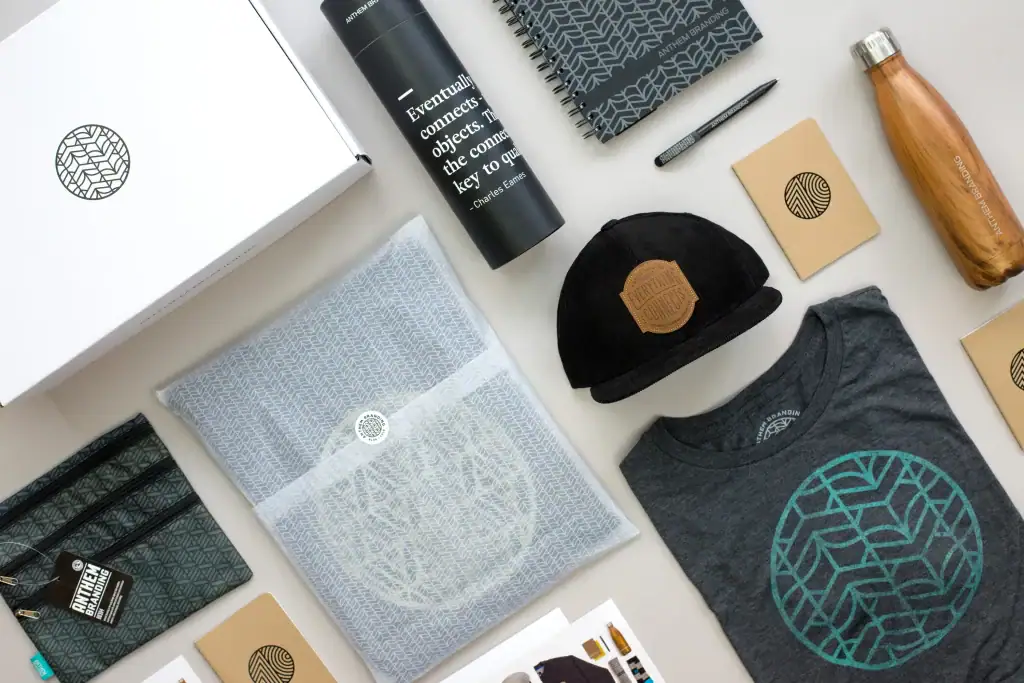 Flat lay of branding goodies: black tumbler, geometric notebooks, cap with leather patch, teal logo t‑shirt, and boxed items arranged on a light surface.