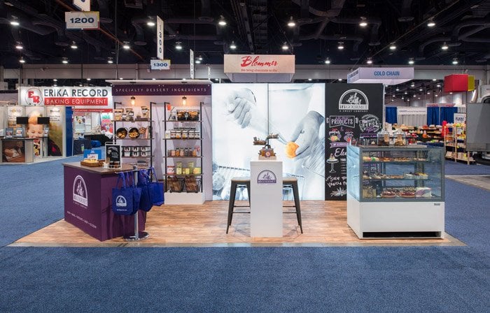 Trade show booth with Blommer signage, a small demonstration press on a white pedestal in the center, a refrigerated display case to the right, and product shelves to the left on a blue carpeted trade hall floor.