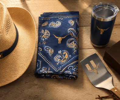 Western themed promotional products flat-lay including a straw cowboy hat, navy paisley bandana, stainless steel BBQ flipper, branded tumbler, and leather gift box on rustic wood surface