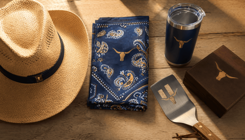 Western themed promotional products flat-lay including a straw cowboy hat, navy paisley bandana, stainless steel BBQ flipper, branded tumbler, and leather gift box on rustic wood surface