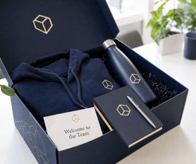 Open custom-branded corporate welcome kit box containing a navy hoodie, insulated water bottle, leather notebook, pen, and welcome card on a white desk